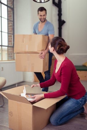 Happy young couple moving into a new home smiling at each other as the wife unpacks a carton on the floor as the husband arrives with two more